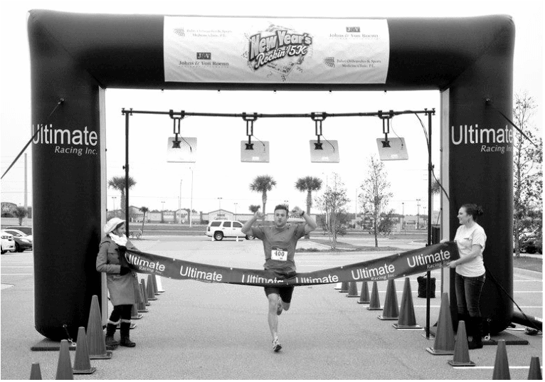 Runner breaking the finish tape under the Ultimate Racing arch with overhead RFID chip timing antennas at a Florida 5K